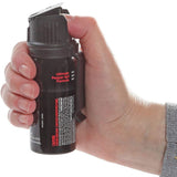 Close view of a person gripping the Mace Magnum 3 pepper gel canister with fingers wrapped around the body and thumb positioned to lift the flip‑top.