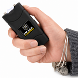 A hand holding a black pet stun gun with a visible LED light on the front, featuring the 'Smart Vet' and 'SMACK PRODOG' branding. The hand is wearing a gray knit sweater and silver bangles with a dog tag charm.